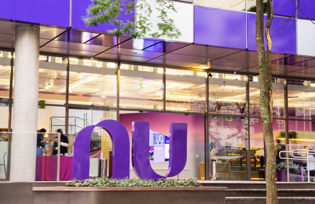 Fachada moderna roxa e de vidro com letras gigantes 'nu' na frente. Pessoas são vistas dentro do prédio.