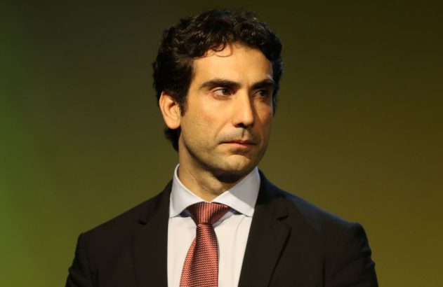 Retrato de homem de terno, gravata e cabelo escuro cacheado, olhando para a esquerda.