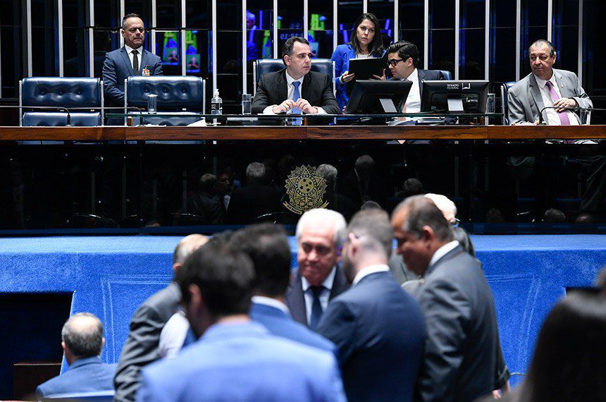 Sessão parlamentar: políticos interagem no plenário; outros estão sentados em mesa diretora.