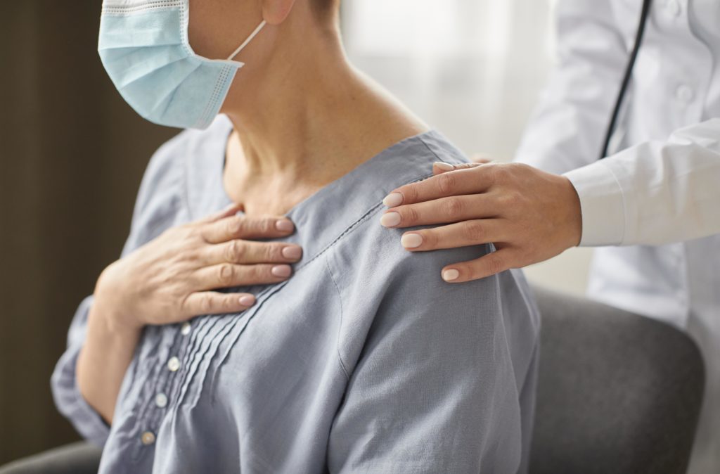 Paciente com máscara e mão no peito recebe apoio de médica(o) em consulta.