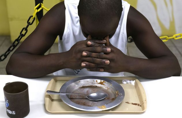 Homem com cabeça baixa e mãos unidas sobre prato com restos de comida.