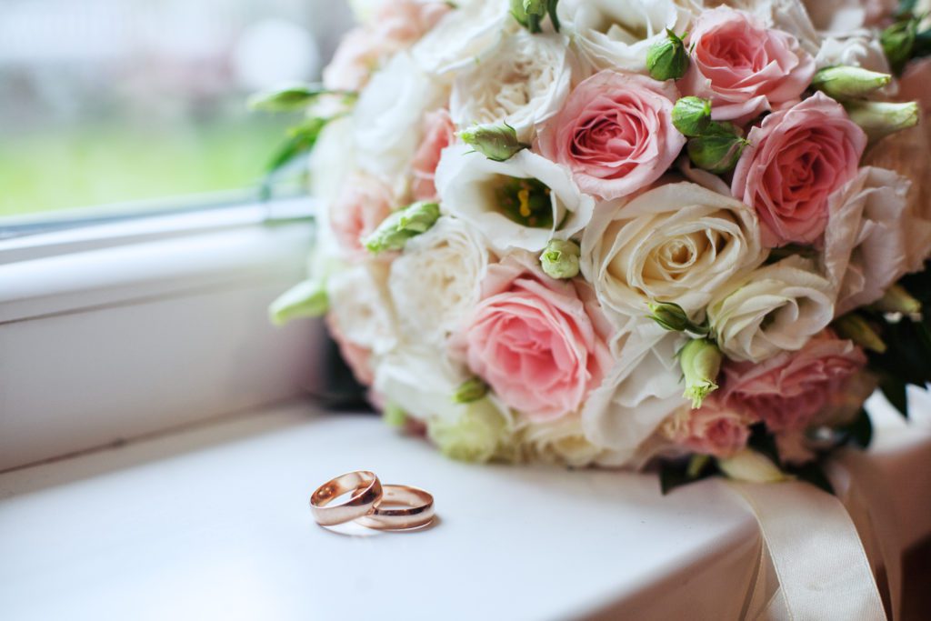 Buquê de casamento com rosas cor-de-rosa e brancas ao lado de dois anéis dourados sobre um peitoril branco.
