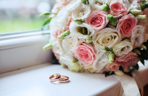 Buquê de casamento com rosas cor-de-rosa e brancas ao lado de dois anéis dourados sobre um peitoril branco.