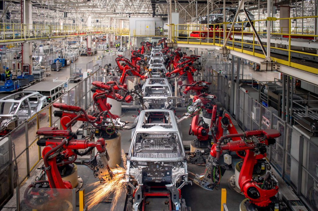 Fábrica de carros: robôs vermelhos montam carrocerias em linha de produção. Faíscas de soldagem em primeiro plano.