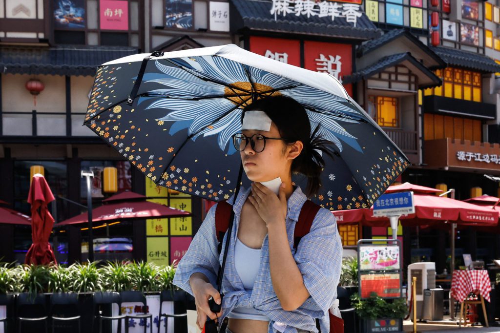 Jovem com adesivo na testa usa sombrinha florida para se proteger do sol em rua com arquitetura chinesa.