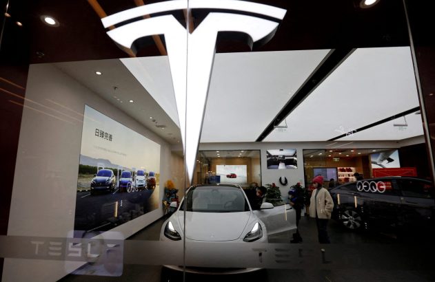 Showroom Tesla com carro branco em exibição, grande logo iluminado da marca e clientes.