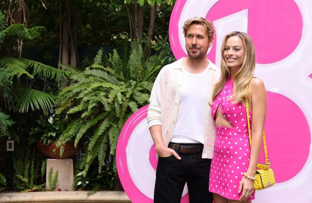 Atores Ryan Gosling e Margot Robbie sorriem posando para o filme Barbie, com fundo rosa e folhagem.