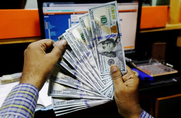 Mãos segurando um leque de notas de 100 dólares americanos em ambiente financeiro.