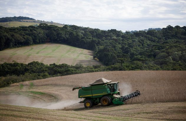 Colheitadeira verde colhendo lavoura dourada em campo inclinado, levantando poeira. Florestas e colinas verdejantes ao fundo.