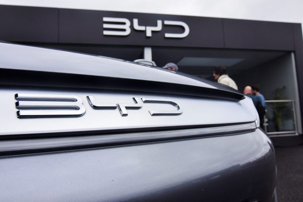 Carro BYD cinza com emblema na traseira e logo BYD grande em concessionária ao fundo.