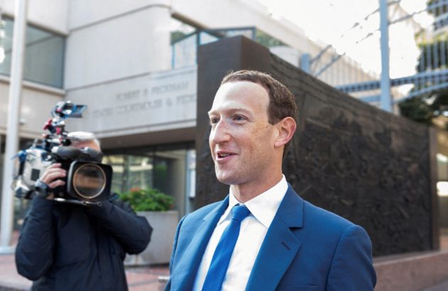 Mark Zuckerberg de terno azul caminhando e sorrindo, com cinegrafista e prédio ao fundo.