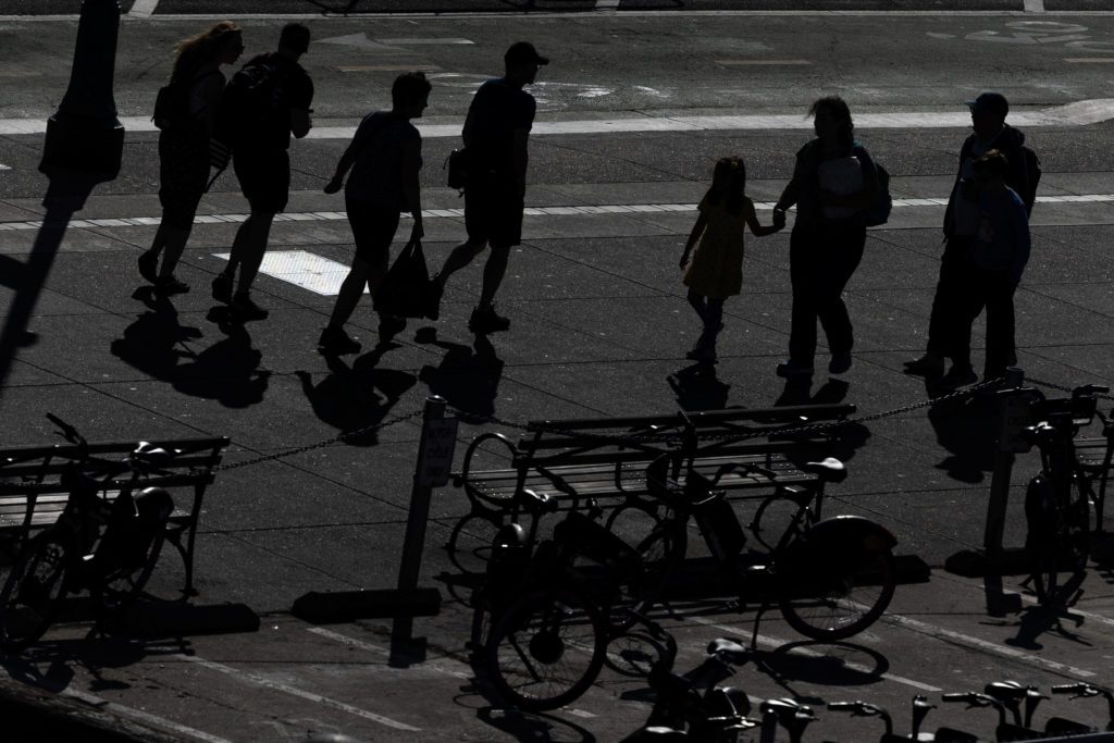 Silhuetas de pessoas caminhando, incluindo criança, em calçada ensolarada. Bicicletas e sombras longas visíveis.