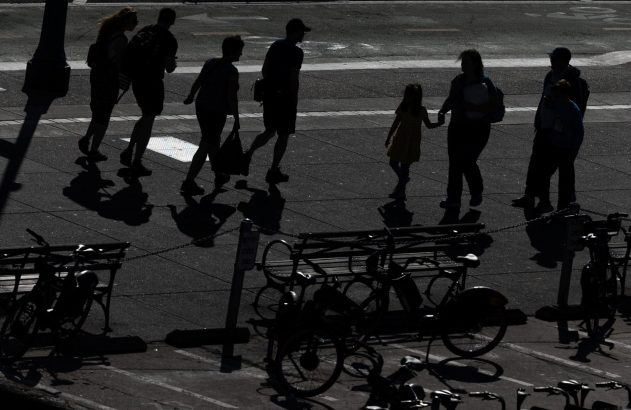 Silhuetas de pessoas caminhando, incluindo criança, em calçada ensolarada. Bicicletas e sombras longas visíveis.