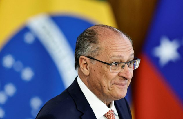 Geraldo Alckmin de terno e óculos, sorrindo. Bandeiras do Brasil e de outro país desfocadas ao fundo.