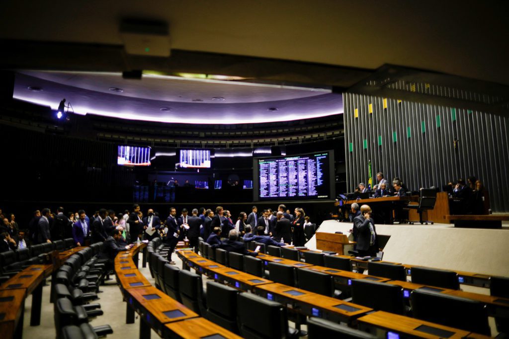 Sessão em plenário legislativo com pessoas sentadas em assentos curvos e telões modernos.