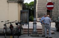Dois homens conversam em rua urbana. Bicicletas, poste de luz e placa de "sentido proibido" à vista.