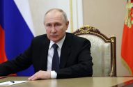 Vladimir Putin sentado à mesa, com bandeiras russa e presidencial ao fundo, olhando para frente.