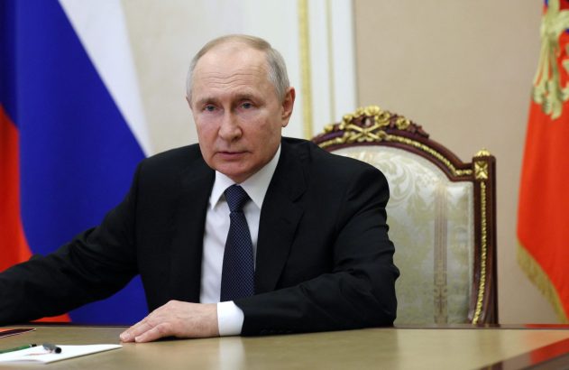 Vladimir Putin sentado à mesa, com bandeiras russa e presidencial ao fundo, olhando para frente.