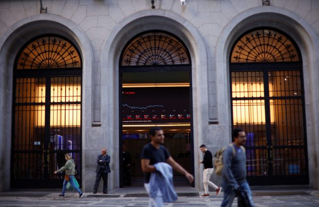 Prédio com arcos e tela de cotações da bolsa. Pessoas caminham na rua.