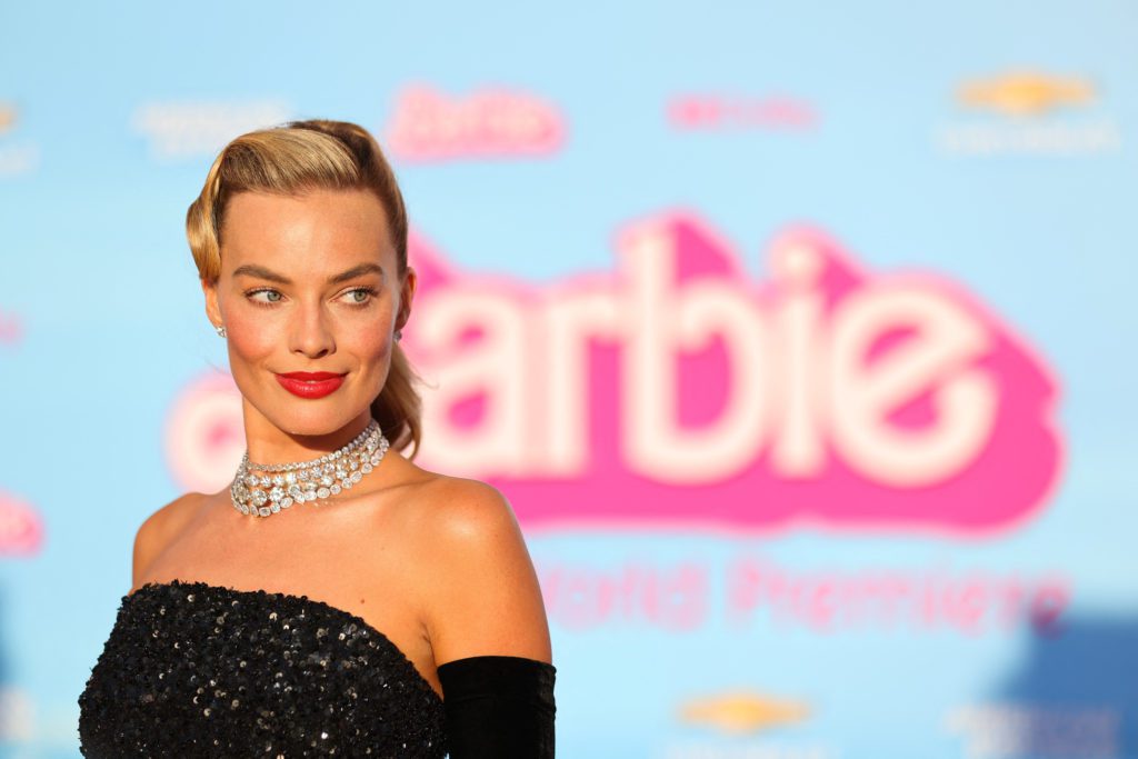Margot Robbie, em vestido preto e colar de diamantes, sorri na estreia do filme Barbie. Fundo azul com logo rosa.