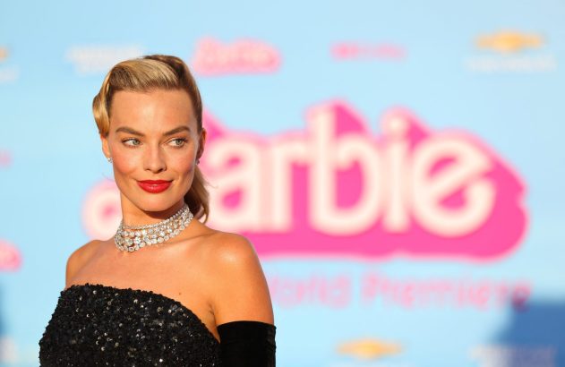 Margot Robbie, em vestido preto e colar de diamantes, sorri na estreia do filme Barbie. Fundo azul com logo rosa.