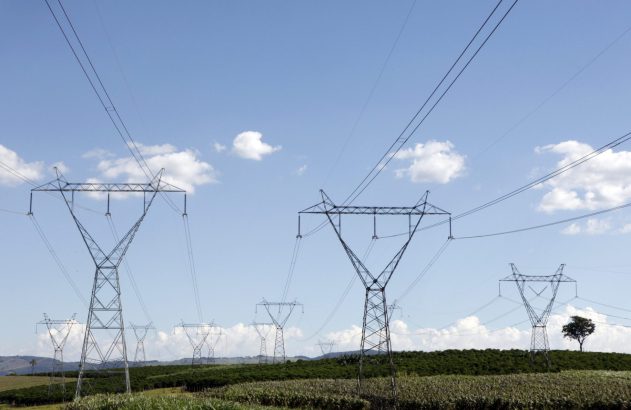 Linhas de transmissão de energia com várias torres metálicas em campo verde sob céu azul.