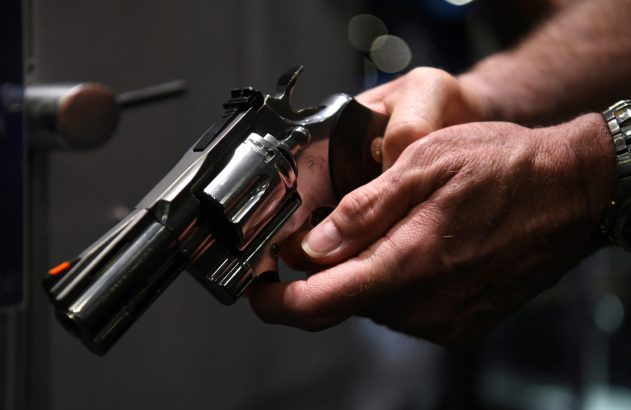 Mãos seguram um revólver com acabamento brilhante, visto em detalhe.