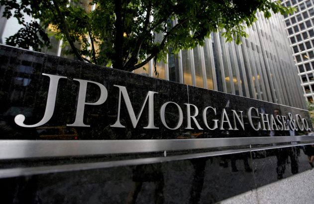 Placa preta polida com "JPMORGAN CHASE & Co." em letras prateadas, reflexos de pessoas, edifício e árvore.
