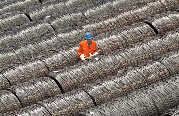 Trabalhador de capacete e laranja inspeciona grandes bobinas de arame de metal em fileiras.