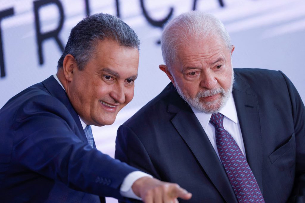 Um homem sorri e aponta, enquanto o presidente Lula, ao seu lado, observa atentamente. Ambos vestem terno.