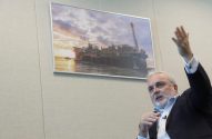 Homem de terno gesticula. Atrás, foto de plataforma petrolífera em mar ao pôr do sol.