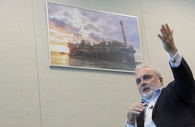 Homem de terno gesticula. Atrás, foto de plataforma petrolífera em mar ao pôr do sol.