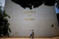 Dois homens caminham à frente do Ministério da Fazenda. O nome do ministério está escrito em letras grandes na fachada.