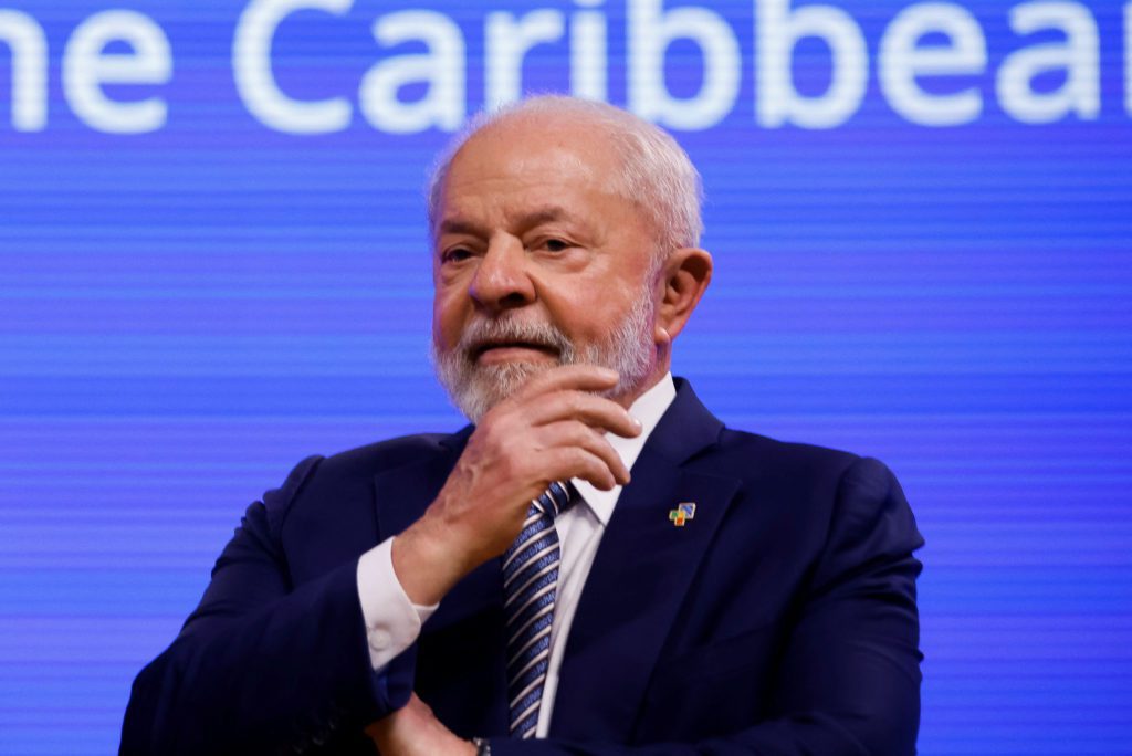 Homem de terno azul, cabelo e barba brancos, com a mão no queixo. Fundo azul com o texto 'e Caribbean'.