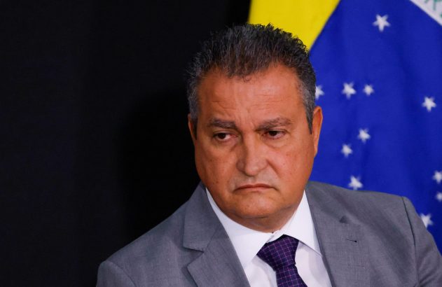 Homem de terno cinza e gravata, com a bandeira do Brasil ao fundo.