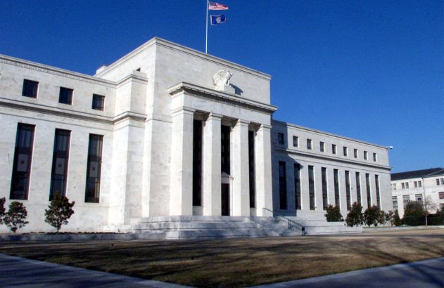 Edifício do Federal Reserve dos EUA com duas bandeiras hasteadas. Águia esculpida sobre colunas, sob céu azul claro.