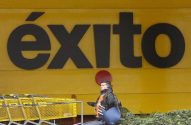 Homem agachado à frente do letreiro ÉXITO (preto em fundo amarelo), com carrinhos de supermercado amarelos.