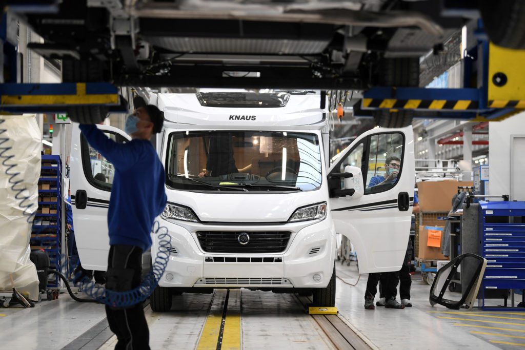 Trabalhador na linha de montagem inspeciona a parte inferior de um motorhome, com outros veículos sendo montados.