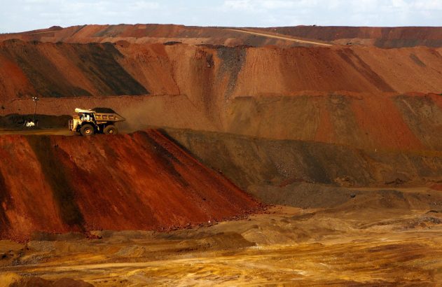 Caminhão de mineração carregado em vasta mina a céu aberto, com terreno em camadas de tons vermelhos e marrons.