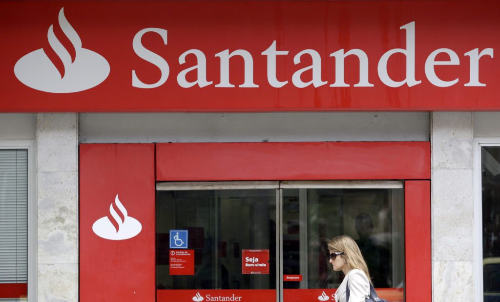 Fachada vermelha do Banco Santander com logotipo e uma mulher passando em frente à agência.