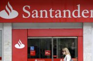 Fachada vermelha do Banco Santander com logotipo e uma mulher passando em frente à agência.