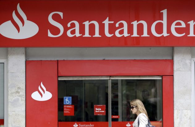 Fachada vermelha do Banco Santander com logotipo e uma mulher passando em frente à agência.
