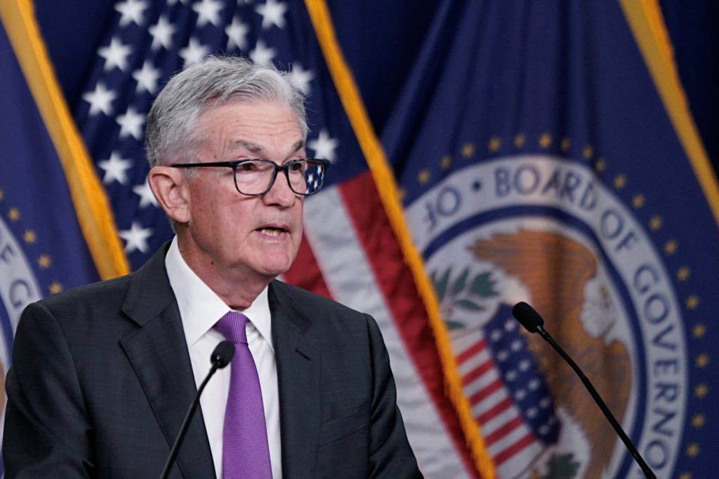 Homem de terno e óculos discursa em pódio com microfone, diante de bandeiras dos EUA e do Federal Reserve.