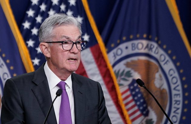 Homem de terno e óculos discursa em pódio com microfone, diante de bandeiras dos EUA e do Federal Reserve.