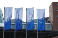 Quatro bandeiras azuis da UE com estrelas amarelas, alinhadas em frente a um edifício moderno.