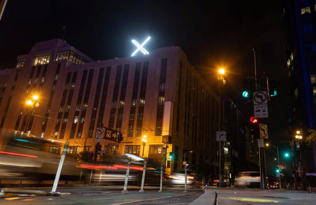 Prédio com 'X' luminoso à noite, com rastros de luz de veículos na rua.