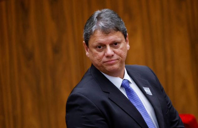 Um homem de terno escuro e gravata azul, olhando levemente para o lado. Fundo amadeirado.