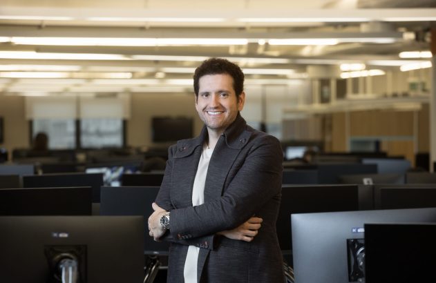 Homem de braços cruzados sorri para a câmera em laboratório de informática.