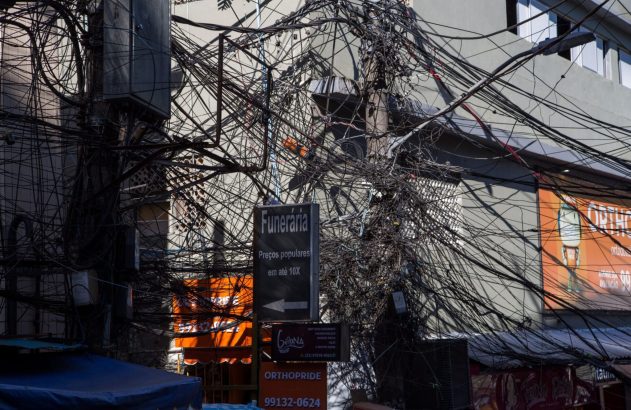Emaranhado denso de fios e cabos em postes de rua urbana, com a placa "Funerária" e outros comércios visíveis.