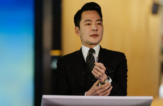 Homem de terno escuro, gravata listrada e lenço de bolso, ajusta o relógio no pulso em estúdio.